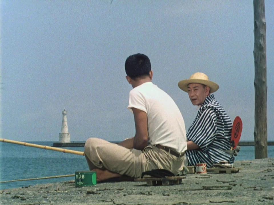 Floating Weeds (1959) movie still: medium — A concrete breakwater extends into the sea under a clear, pale blue sky. Two men sit on the edge of the…