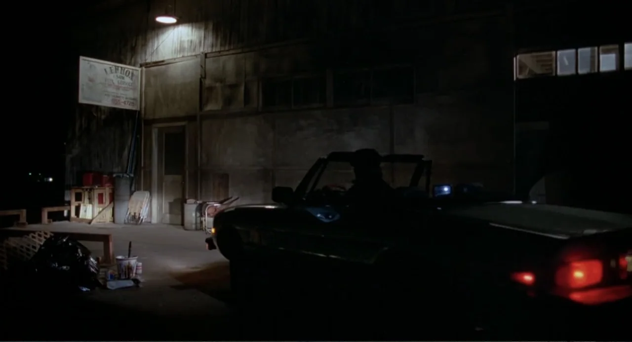 Dangerously Close (1986) movie still: wide — A dark, industrial building exterior at night features a weathered sign and a convertible. A single…