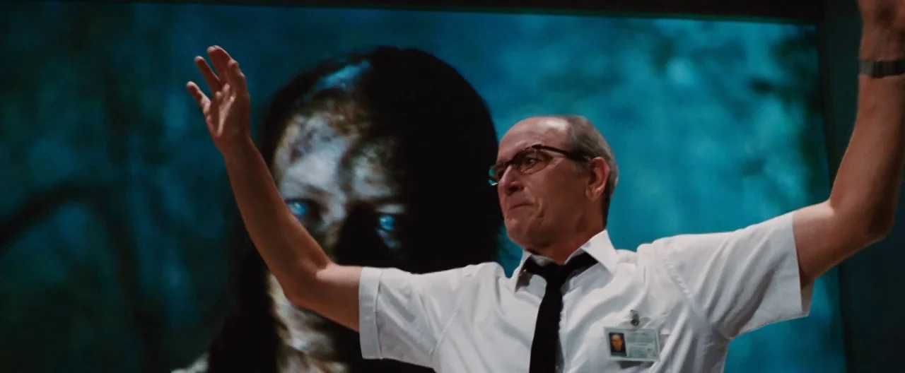 The Cabin In The Woods (2012) movie still: medium — A man in a white shirt stands before a large glowing blue monitor screen. The screen displays a…