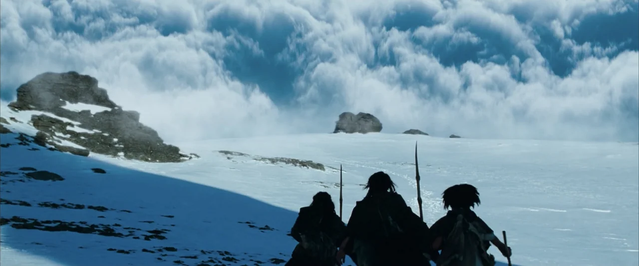 10,000 BC (2008) movie still: wide — Three individuals wearing fur garments trek across a vast, snow-capped mountain ridge under bright sunlight.…
