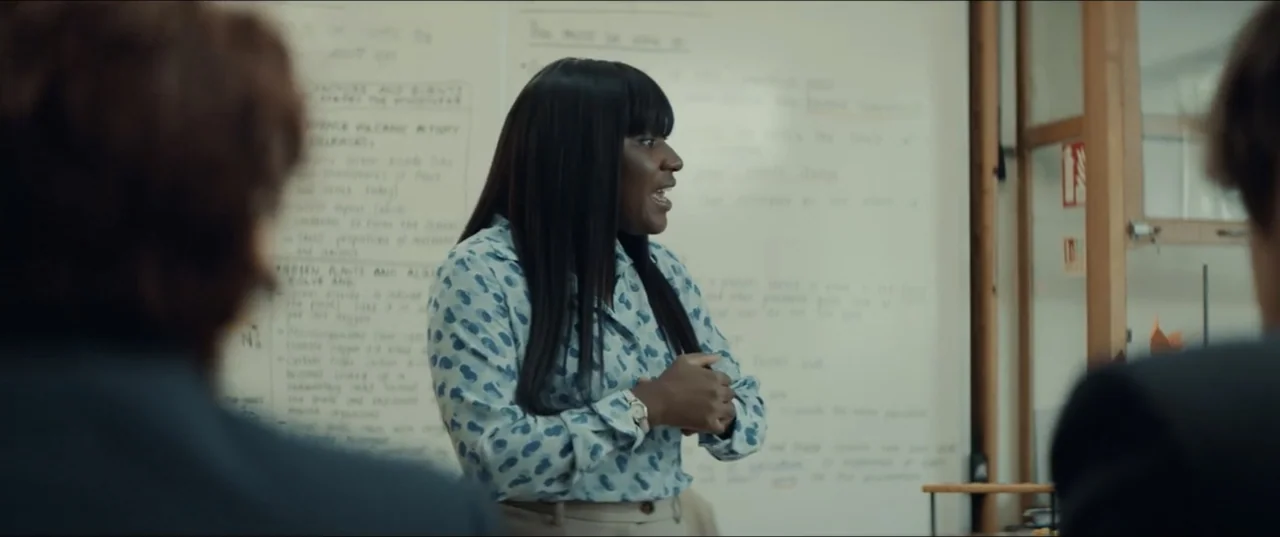 Bonus Track (2024) movie still: medium — A teacher stands before a whiteboard covered in handwritten scientific text and diagrams. A woman wearing a…
