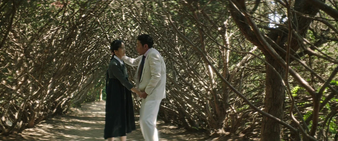 The Handmaiden (2016) movie still: fullbody — A narrow dirt path is enclosed by a dense tunnel of interlocking tree branches. A woman in a…