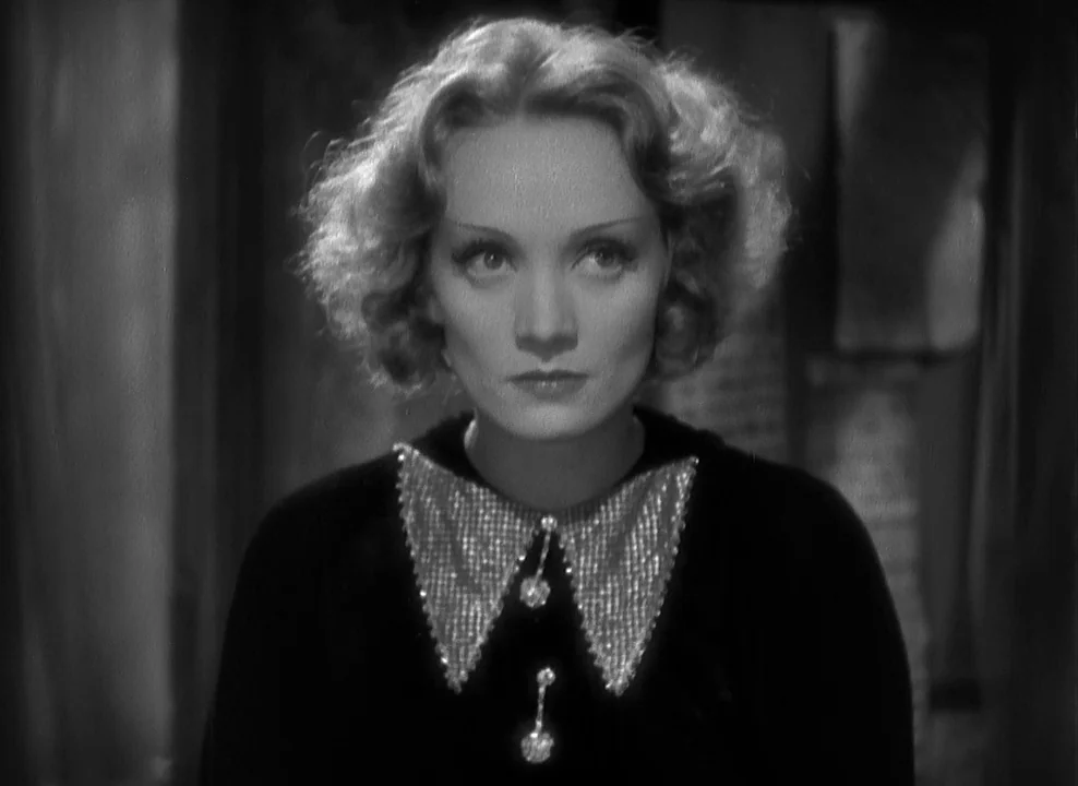 Shanghai Express (1932) movie still: closeup — A woman stands centered against a dark, nondescript interior background. She maintains a steady,…