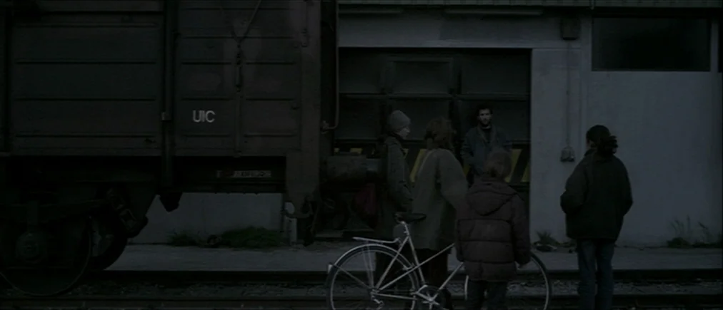 Time Of The Wolf (2003) movie still: wide — An industrial railway yard features a large metal cargo train car and concrete buildings. Four people…