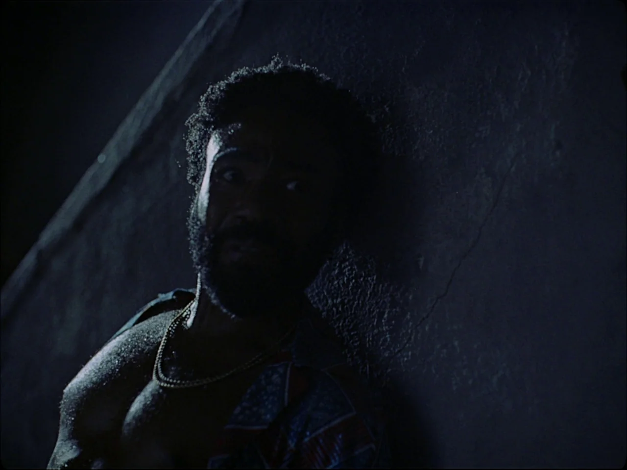 Guava Island (2019) movie still: closeup — A dark, cramped interior with a textured wall surface that catches side lighting. A man with a full beard…
