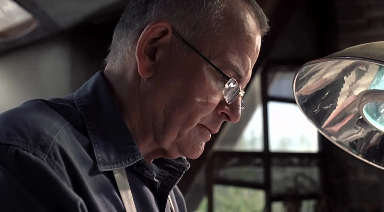 eXistenZ (1999) movie still: closeup — A workshop interior features a man working with focused concentration. The man, an elderly male, peers…