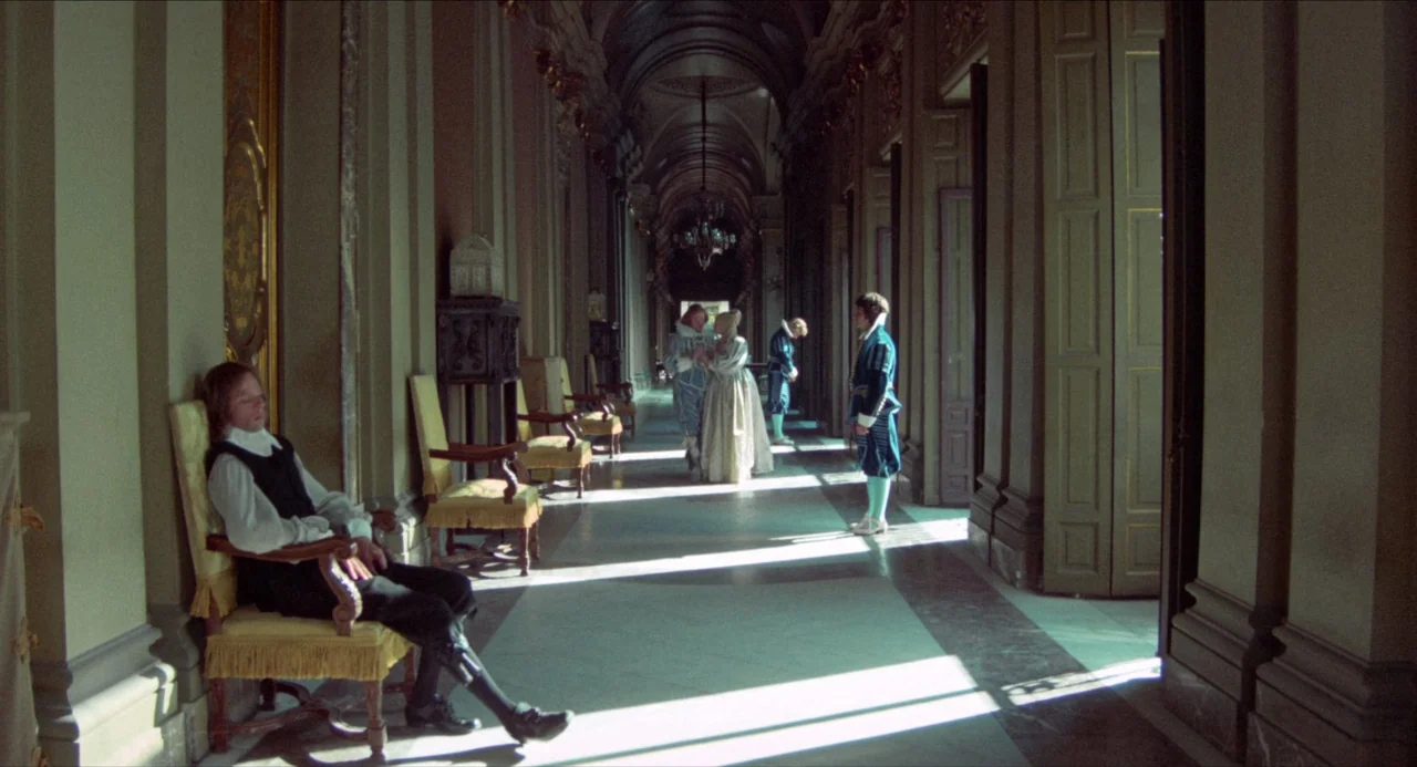 The Three Musketeers (1973) movie still: wide — A long, ornate palace corridor has high ceilings and classical architecture with marble floors. A…