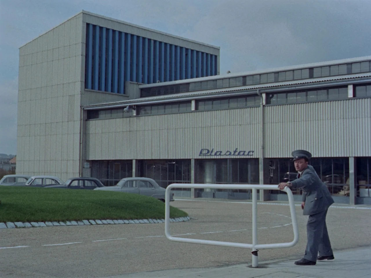 Mon Oncle (1958) movie still: wide — A modern industrial building with 'Plastac' signage stands behind a curved green grass verge. A uniformed…