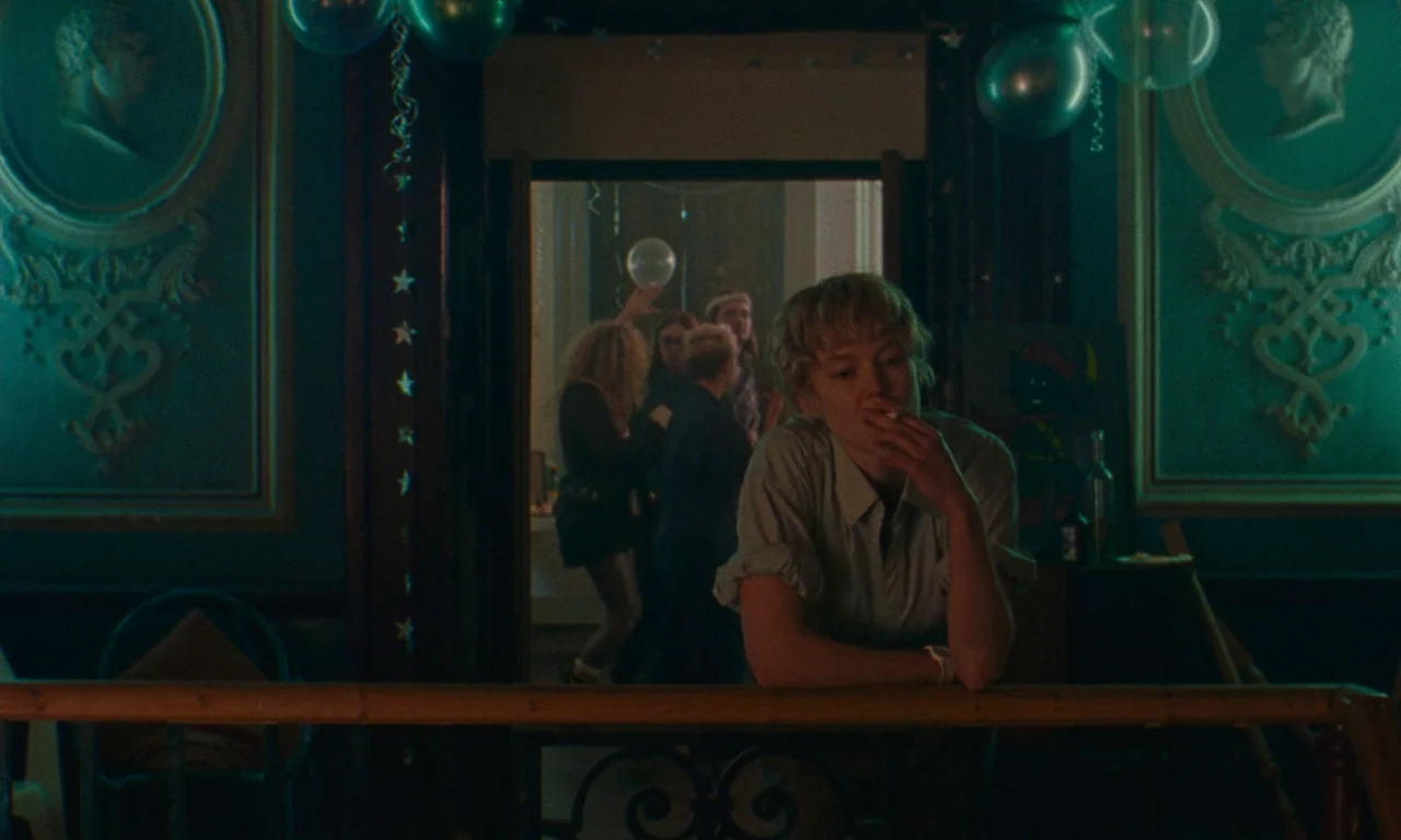 Blue Jean (2022) movie still: medium — A dimly lit room with ornate teal wallpaper features classical cameo wall reliefs. In the foreground, a young…