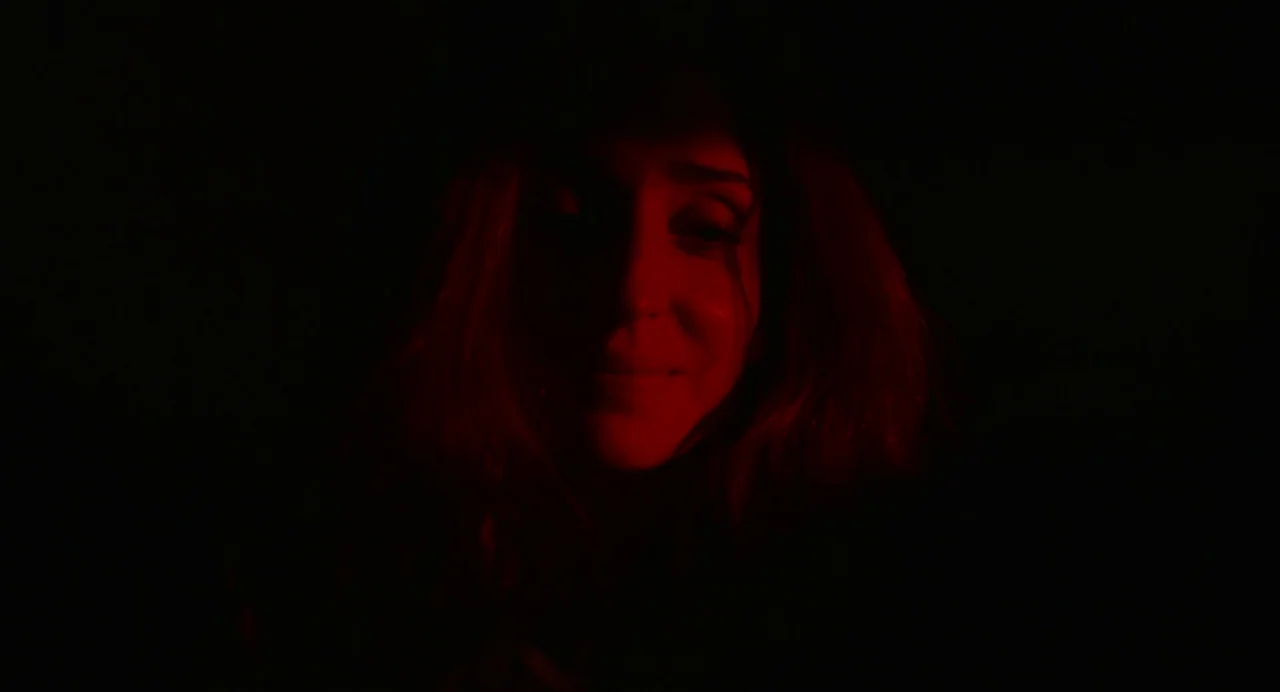 Beau Is Afraid (2023) movie still: closeup — A dark, nondescript interior space completely shrouded in black. A woman's face is partially lit by a…