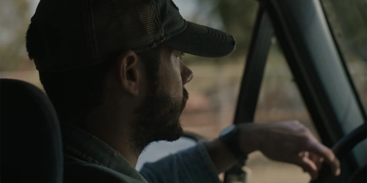 Caddo Lake (2024) movie still: medium — The interior of a vehicle cabin with muted lighting. A man in a baseball cap drives while looking ahead.