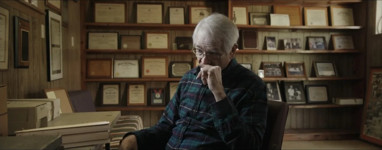 Hunting for Hedonia (2019) movie still: medium — A wood-paneled home office features wall-to-wall shelves filled with framed certificates and…