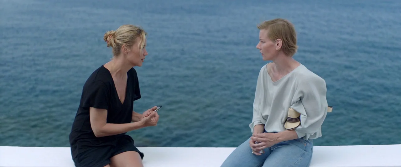 Sibyl (2019) movie still: medium — Two women sit on a white stone wall with the ocean in the background. They are facing each other in conversation.