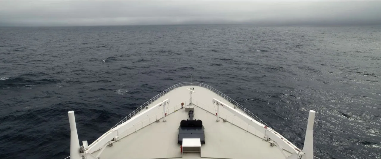 Let Them All Talk movie still: establishing — The pointed bow of a large white ship cuts through a vast, dark ocean. A heavy layer of low grey…