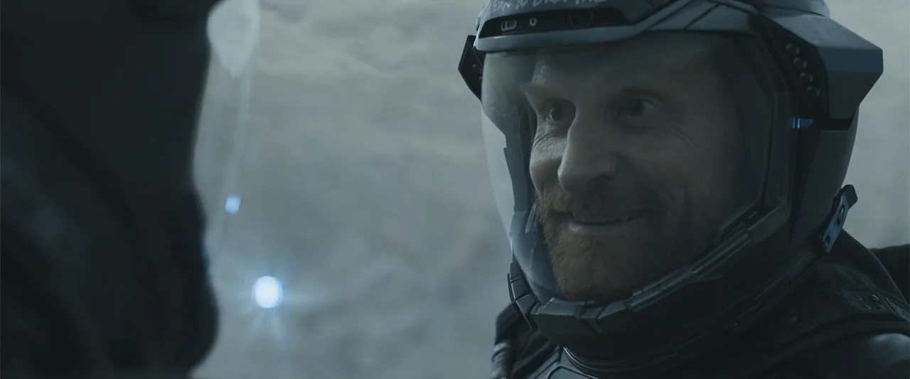 DISTANT (2026) movie still: closeup — A desolate rocky surface at night under dim light conditions. A man wearing a futuristic space helmet looks…