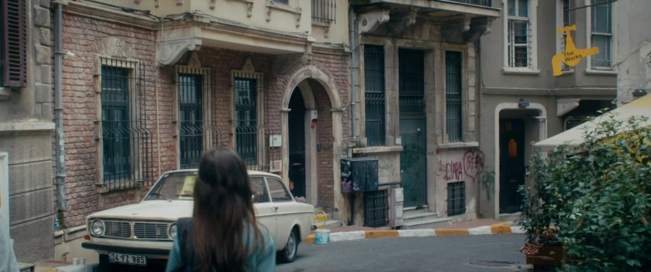 Mustang (2015) movie still: medium — An urban side street features weathered brick buildings with graffiti and a vintage car. A woman with long dark…