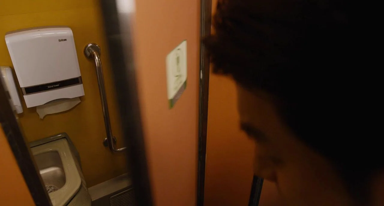 Train To Busan movie still: closeup — A cramped stall in a public restroom features yellow walls and a metal grab rail. A blurred figure stands in…