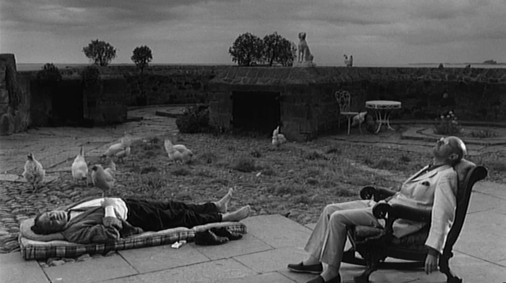 Cul-de-sac (1966) movie still: wide — A stone paved courtyard with a low wall overlooks a body of water. Two men lounge outdoors while several…