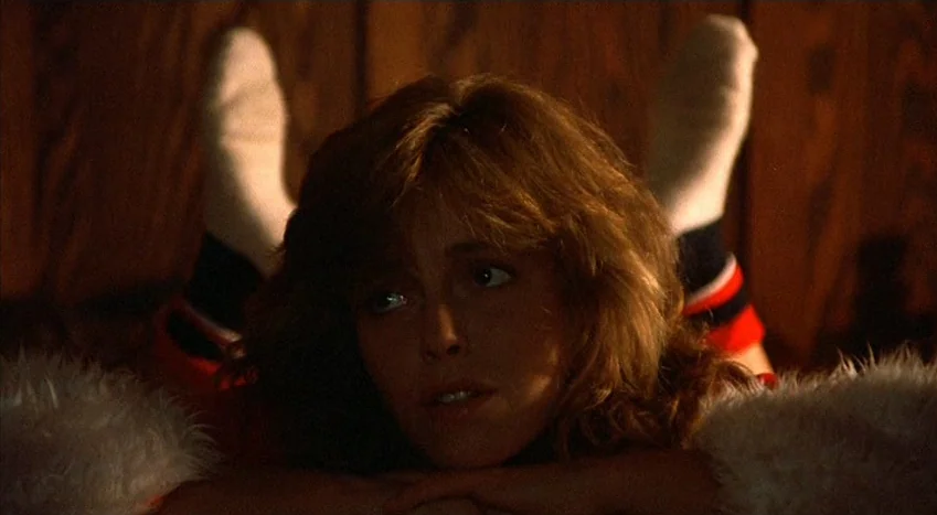 The Coca-Cola Kid (1985) movie still: closeup — A dark, wood-paneled room interior serves as the background for the figure. A young woman lies prone…