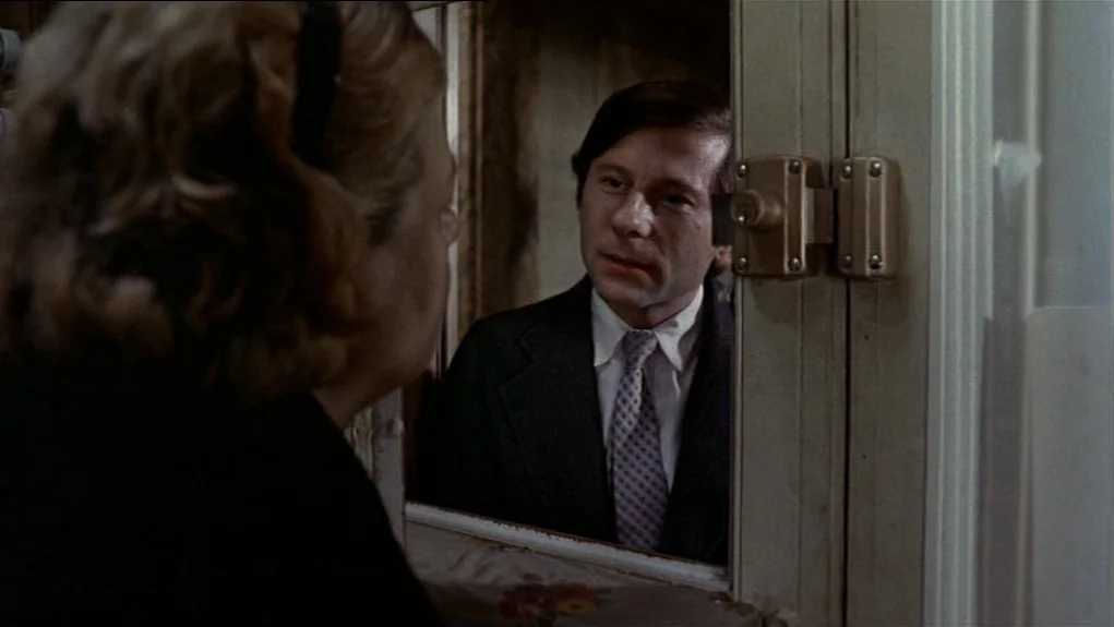 The Tenant (1976) movie still: medium — A woman stands in the foreground viewing a man through a window pane. The man is positioned on the other…