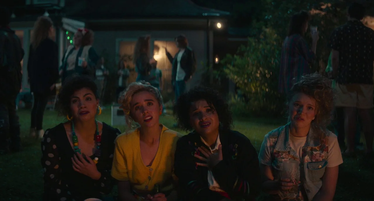 Lisa Frankenstein (2024) movie still: medium — A dark night party takes place on a residential lawn with a house background. Four young women are…