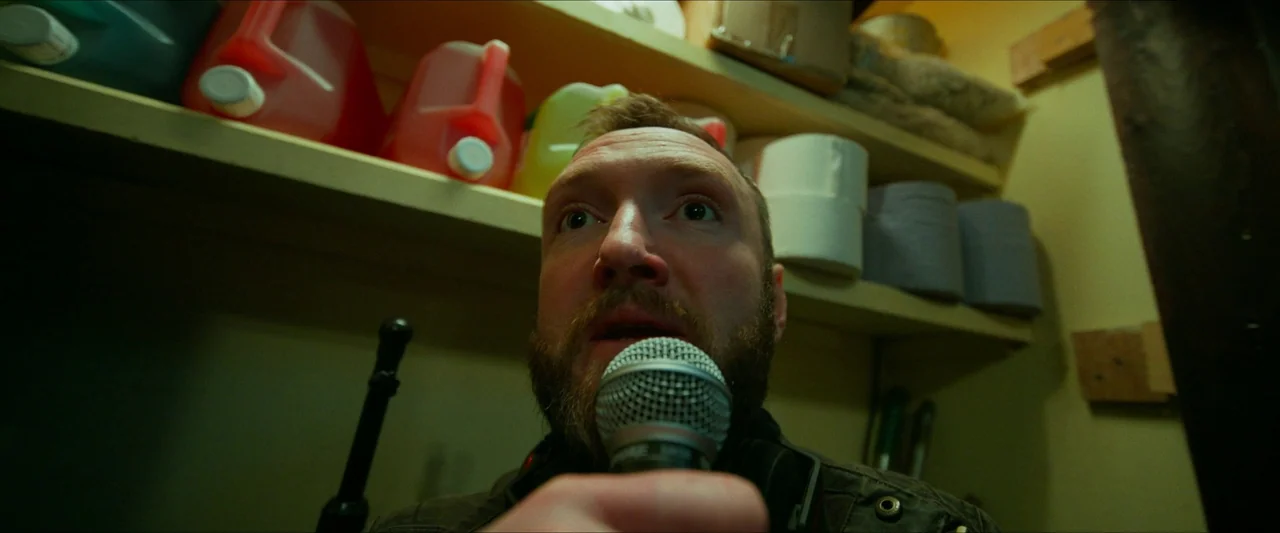 Kneecap (2024) movie still: closeup — A cluttered storage space contains shelves stacked with plastic jugs and paper rolls. A man in a dark jacket…
