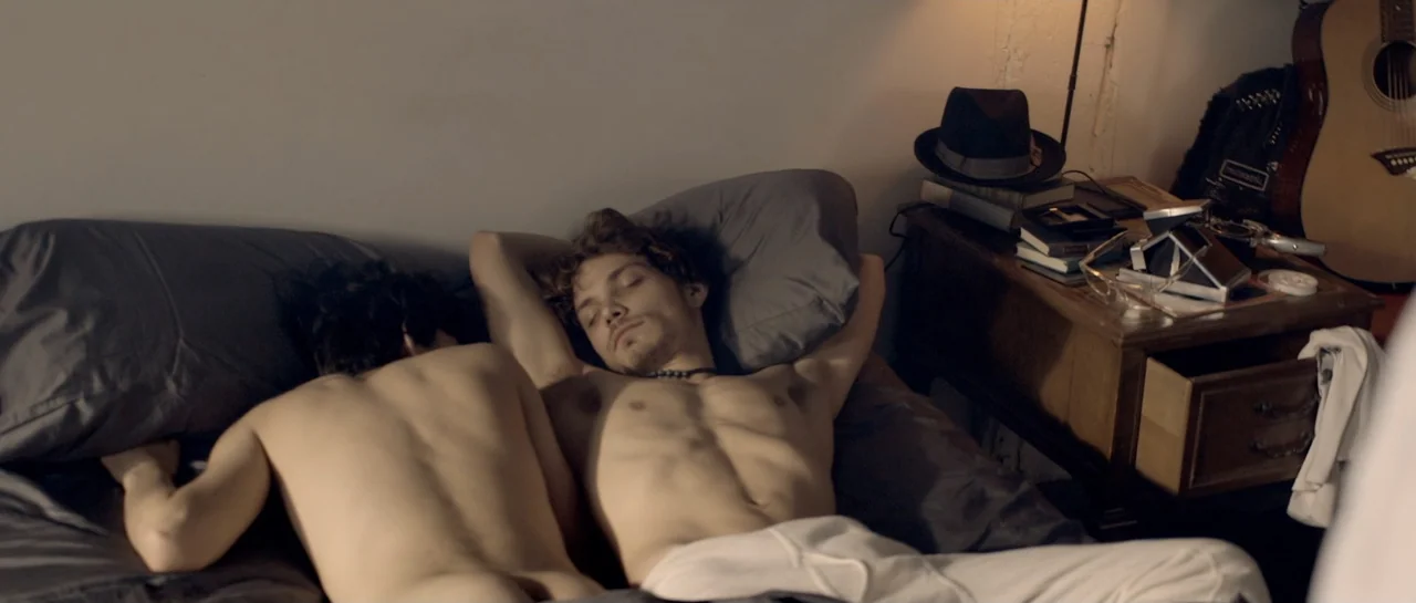 NY84 (2016) movie still: medium — A dimly lit bedroom features a bed with gray linens and a wooden nightstand. Two shirtless young men sleep side by…
