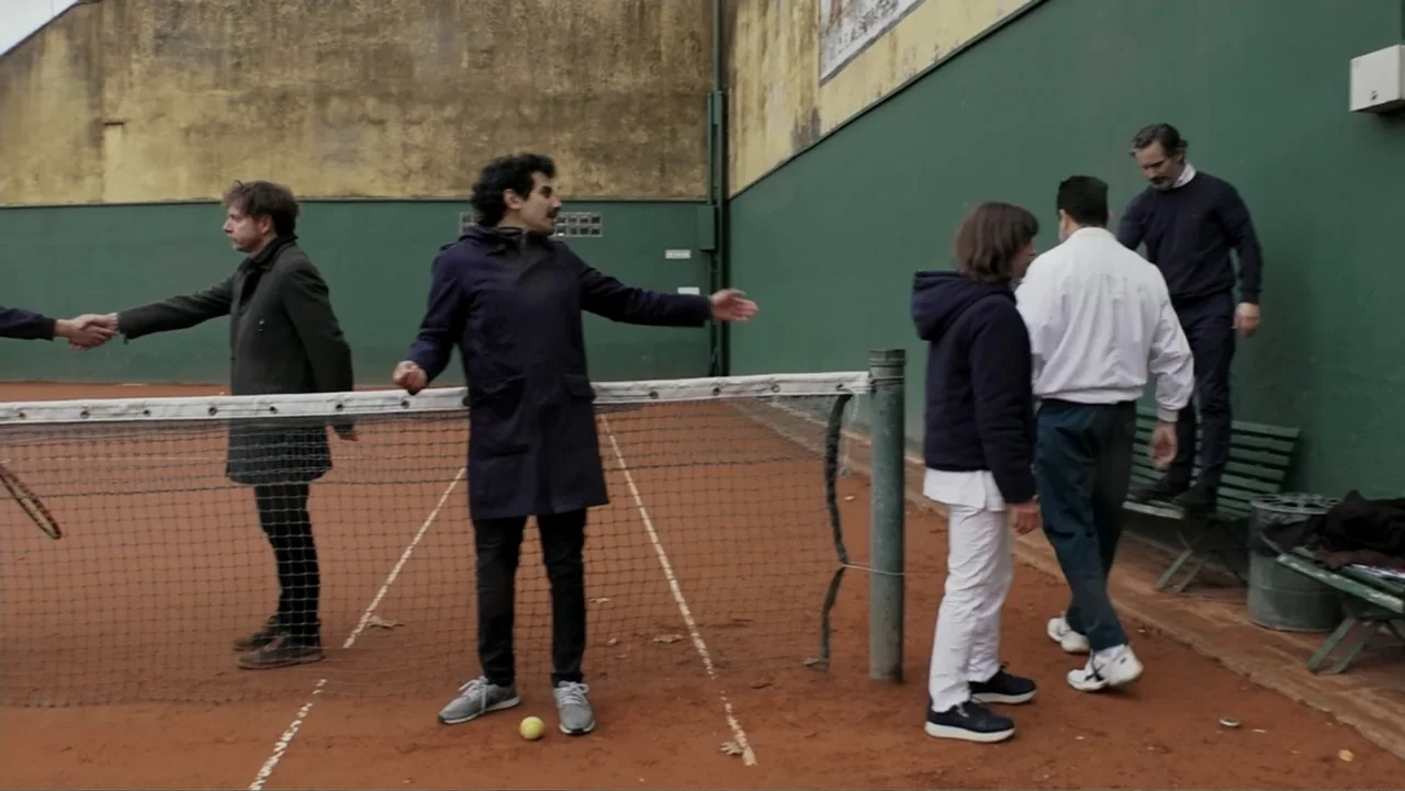 Saturdays Disorders (2022) movie still: fullbody — A worn outdoor clay tennis court features high green perimeter walls and a net. Five people in…