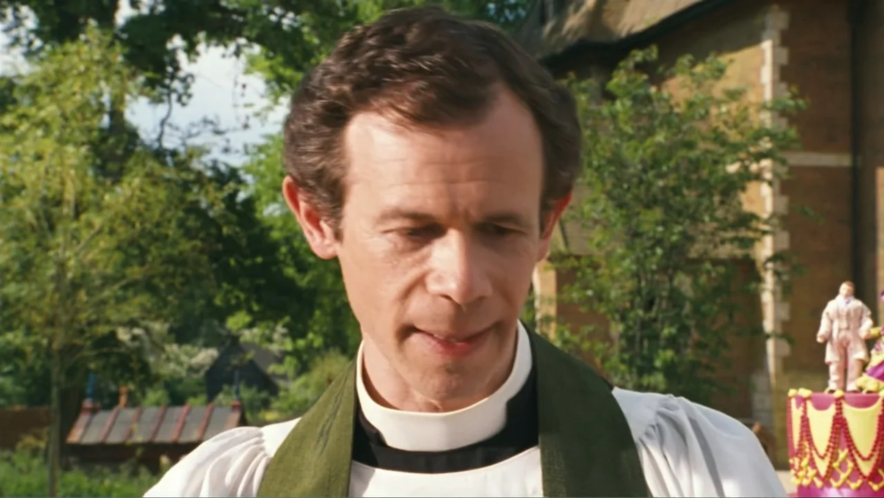 Nanny McPhee and the Big Bang (2010) movie still: closeup — A man in clerical vestments stands outdoors in front of a stone building structure. He…