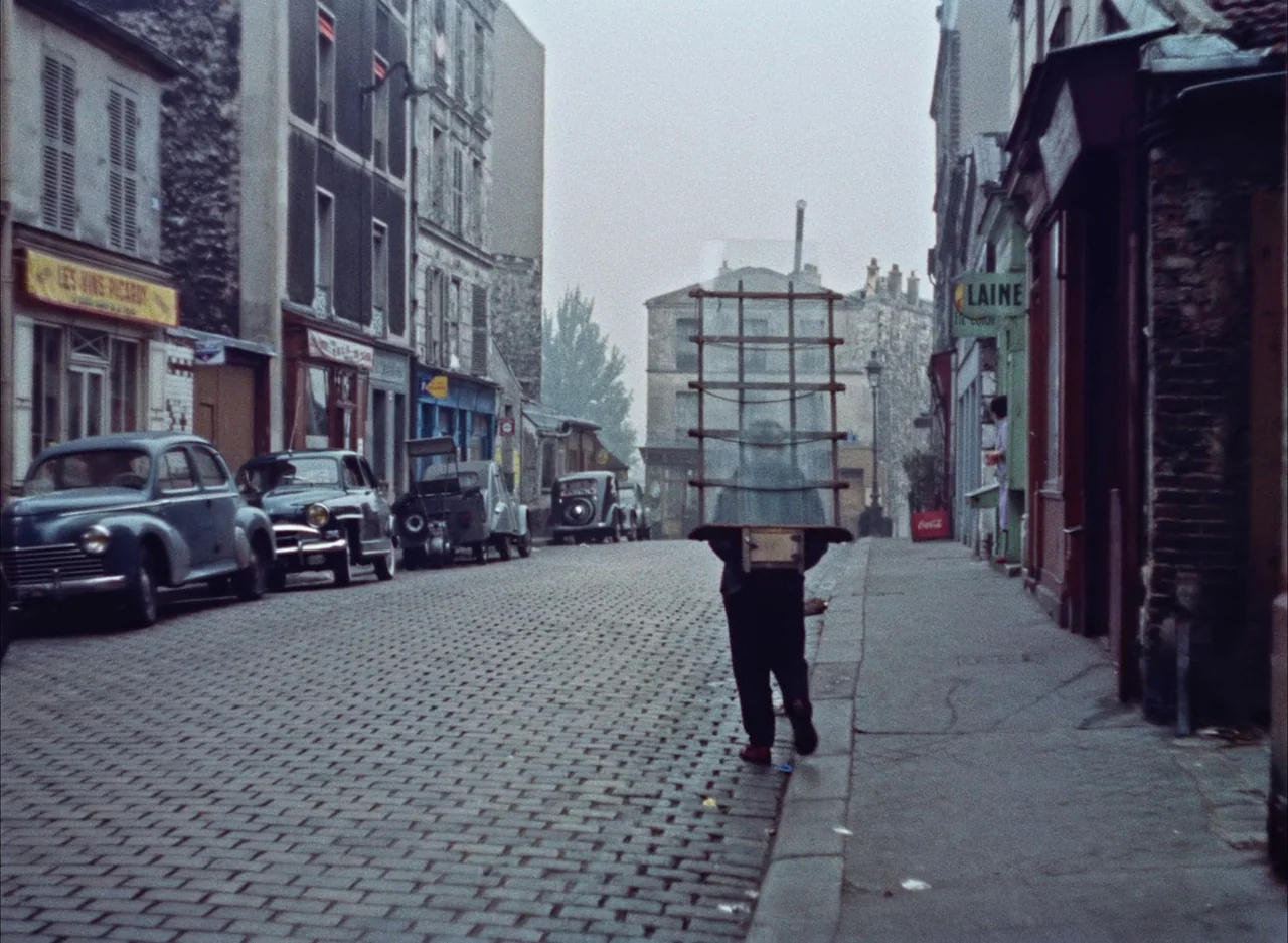 The Red Balloon (1956) movie still: fullbody — A quiet, overcast 1950s European cobblestone street lined with vintage automobiles and shops. A man…