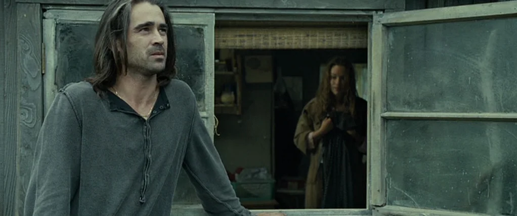 Ondine (2009) movie still: medium — A weathered wooden building features an open window with a person inside. A man stands in the foreground looking…