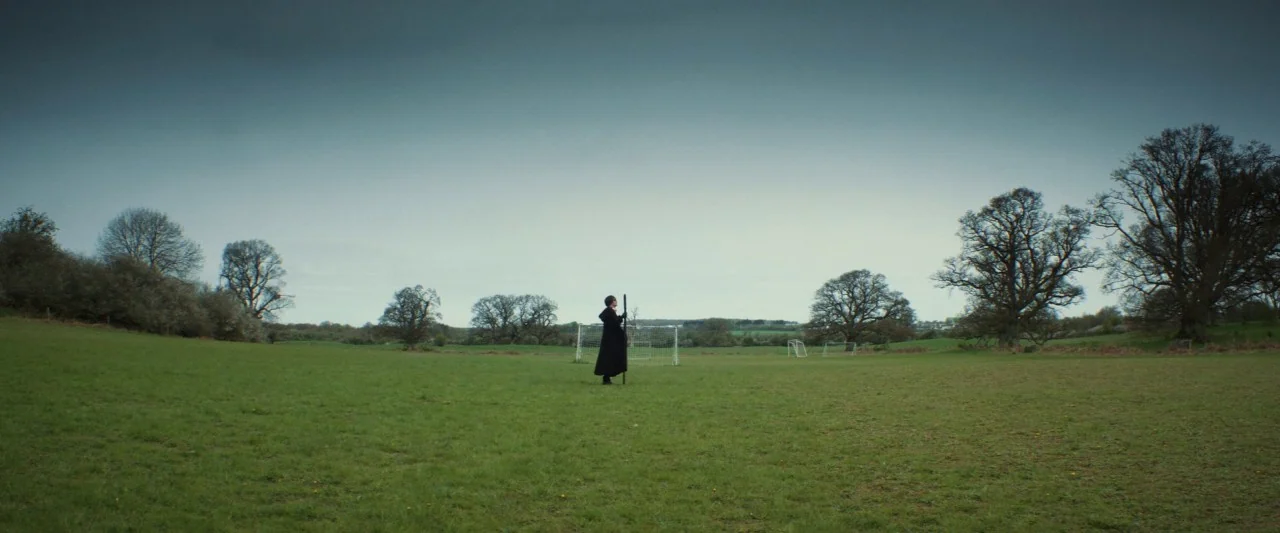 Extra Ordinary (2019) movie still: wide — A vast, open grass field under an overcast, grey sky contains several soccer goals. A lone figure dressed…