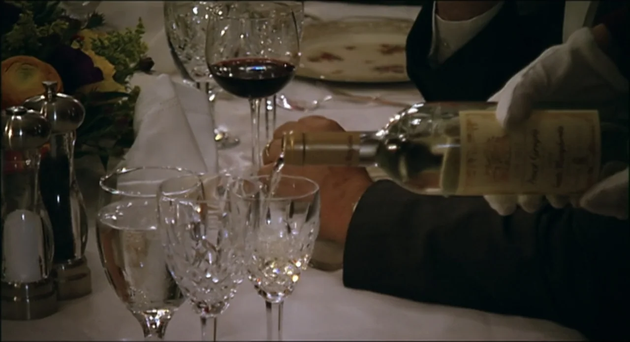 Hard as Nails (2001) movie still: medium — A formal dining table is set with crystal glassware and a wine bottle. A hand wearing white gloves pours…