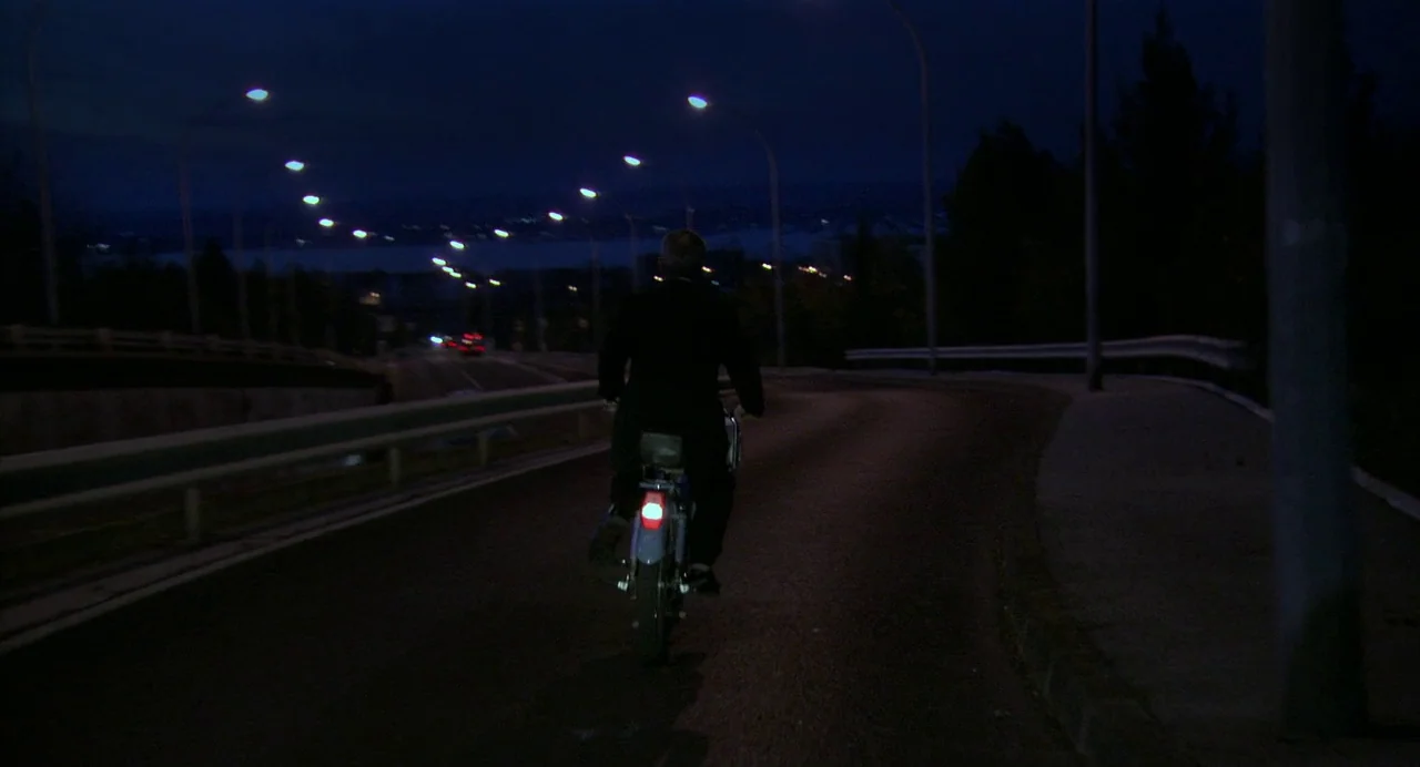 The Secret of the Grain (2007) movie still: wide — A dark, deserted asphalt road stretches forward at night under rows of streetlights. A man rides…