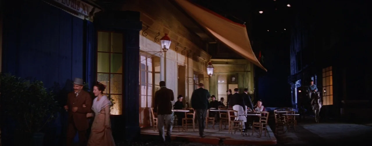 Lust For Life (1956) movie still: wide — A nighttime scene of a 1900s Parisian cafe terrace with wrought iron furniture. A couple walks past the…