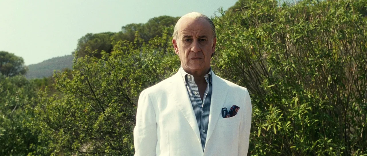 The Great Beauty (2013) movie still: medium — An older man stands before a thick wall of vibrant green leafy bushes. The sunlit outdoor setting…