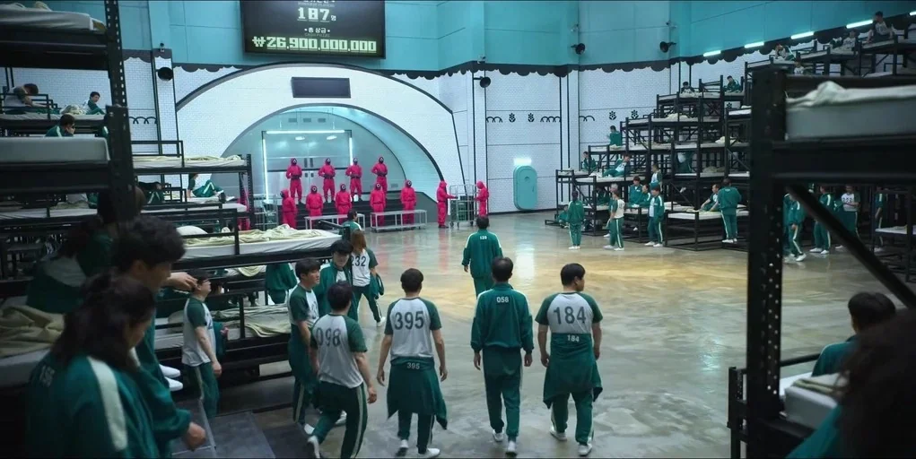 Making of: Squid Game (2021) movie still: wide — A large industrial room features multi-tiered metal bunk beds lining the walls under high ceilings.…