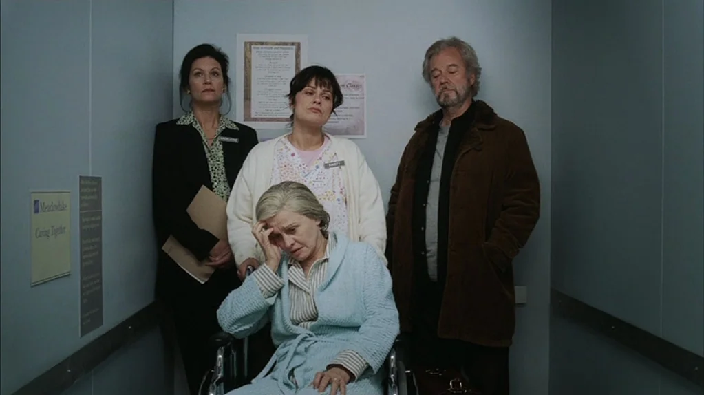 Away From Her (2006) movie still: medium — A small elevator with pale blue walls and posted informational signs. Four adults stand inside, one…