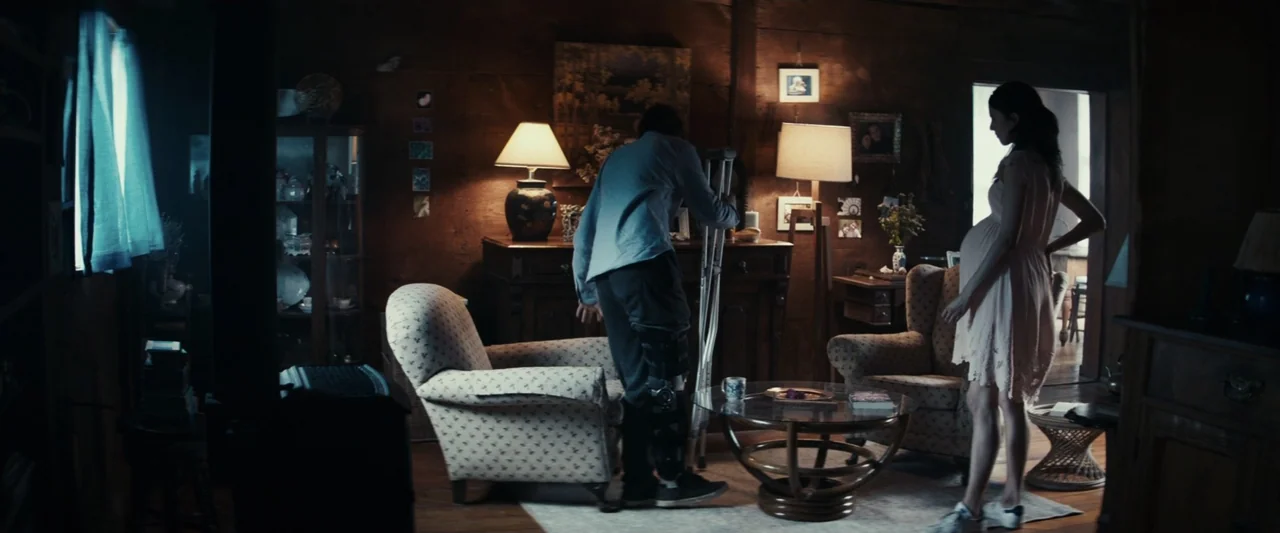 Strange but True (2019) movie still: wide — A rustic wood-paneled living room contains two patterned armchairs and a glass coffee table. A man using…