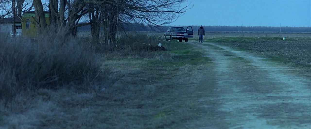 Ballast (2008) movie still: wide — A desolate rural landscape features a dirt path flanked by leafless trees. A man walks forward from a parked…