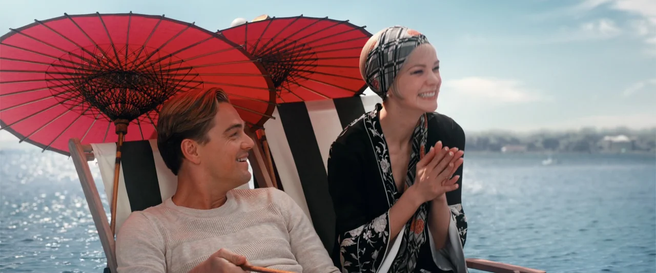 The Great Gatsby (2013) movie still: medium — Two people sit in folding lounge chairs on a boat deck under red parasols. The man and woman smile at…