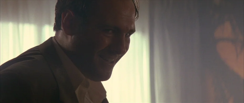 True Romance (1993) movie still: closeup — An interior space is illuminated by bright light streaming through a sheer curtained window. A man with a…