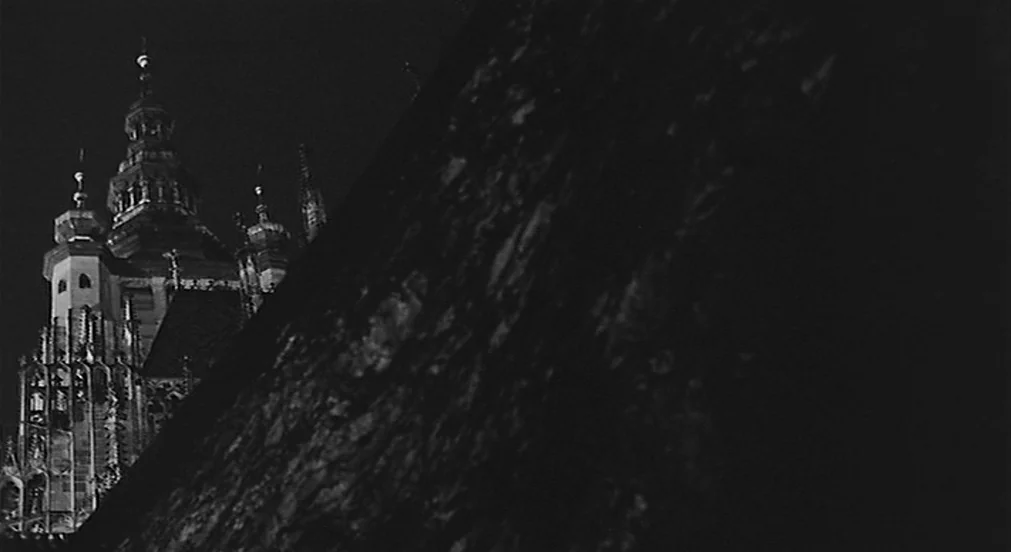 Kafka (1991) movie still: establishing — A dark, massive stone structure occupies the foreground, obscuring part of a gothic cathedral. The…
