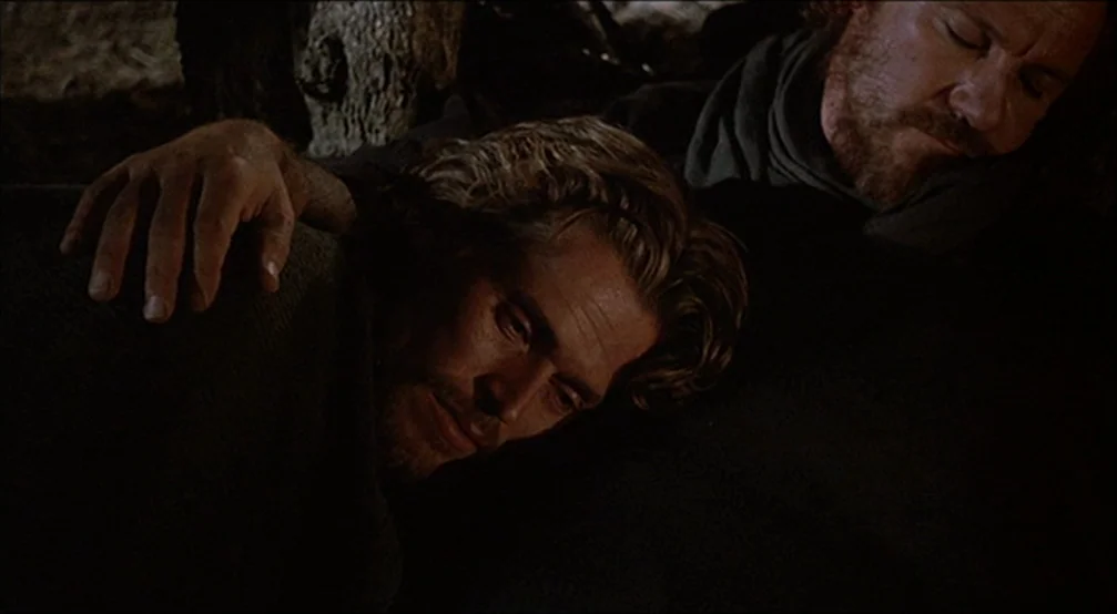 The Last Temptation Of Christ (1988) movie still: closeup — A dark outdoor space with tree bark visible in the background shadow. Two men lie…