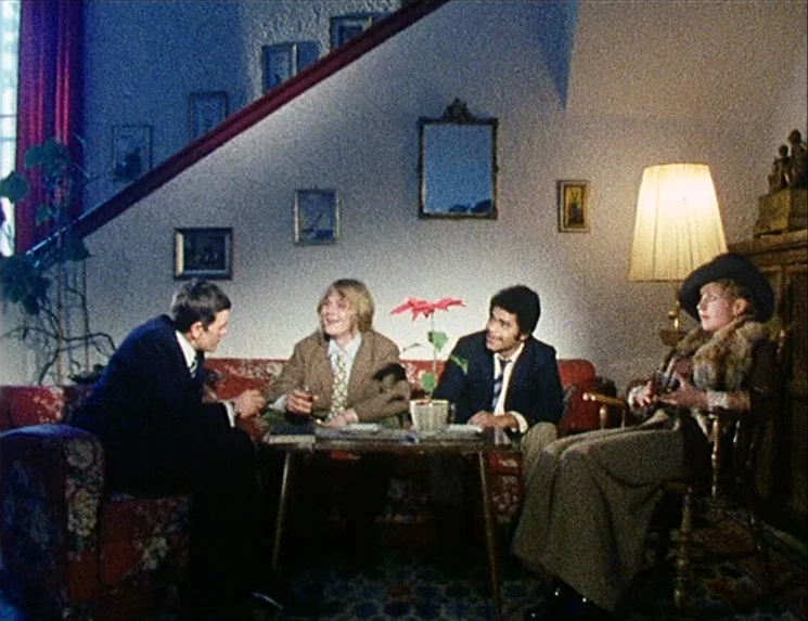 Rio Das Mortes (1971) movie still: wide — A furnished living room features a sofa, a small wooden table, and wall art. Four adults are sitting in…