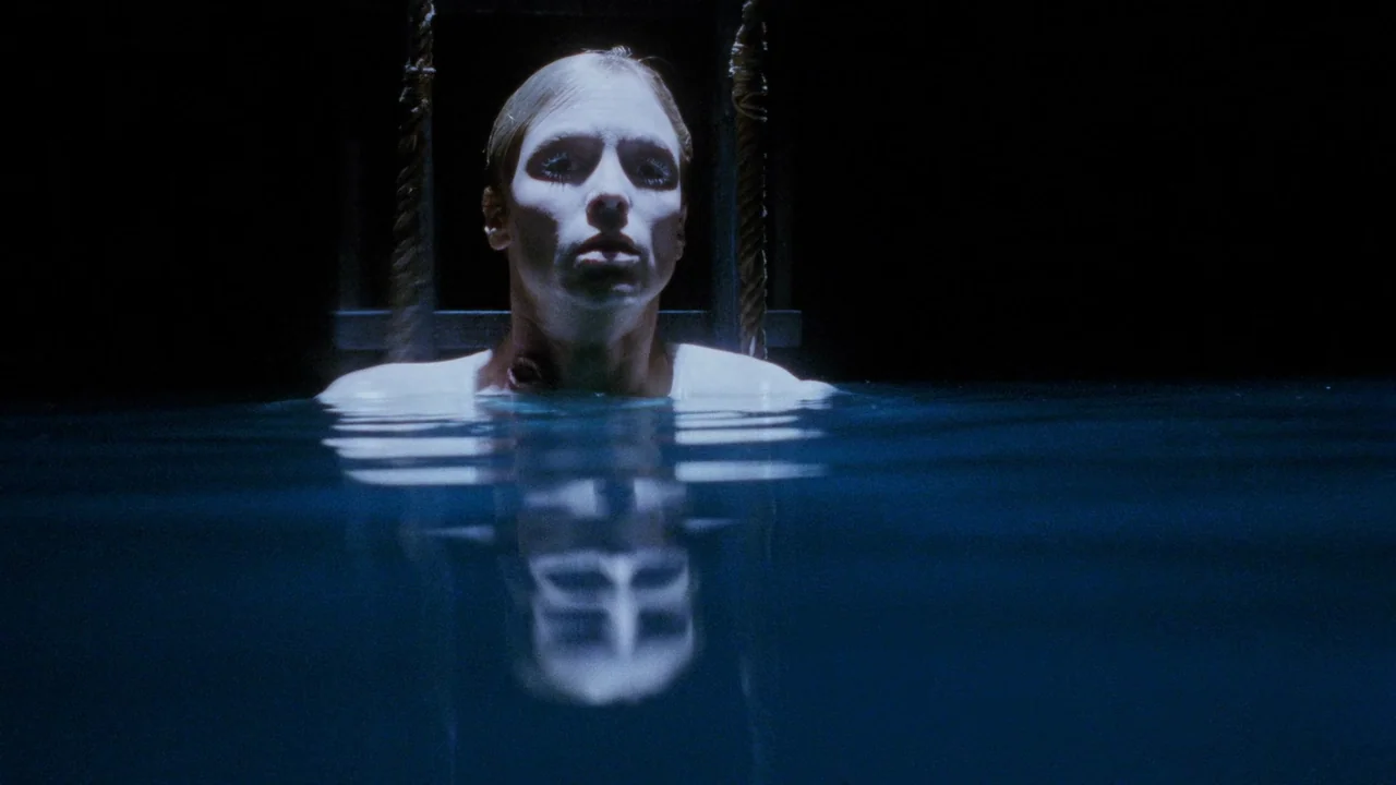 The Sect (1991) movie still: medium — A woman stands in a dark, flooded environment with visible dark water. She stares directly ahead with her…