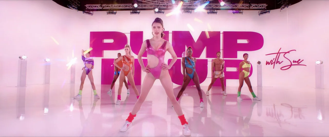 The Substance (2024) movie still: fullbody — A high-key white dance studio features a large magenta logo on the back wall. Seven dancers in colorful…