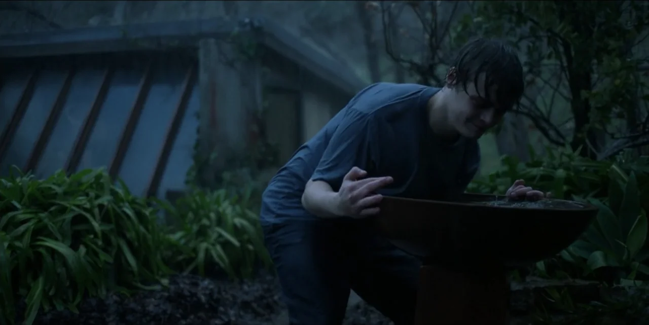 Bring Her Back (2025) movie still: medium — A person stands in a misty outdoor area beside a stone basin and building. A young man wearing a grey…