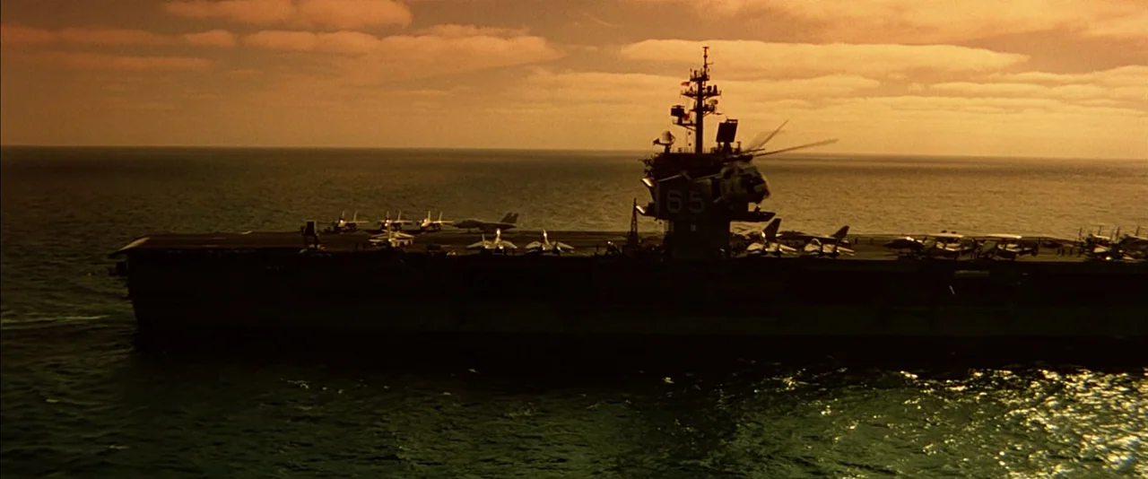 Top Gun (1986) movie still: wide — An aircraft carrier floats on the ocean under a thick layer of golden clouds. Several fighter jets are parked on…