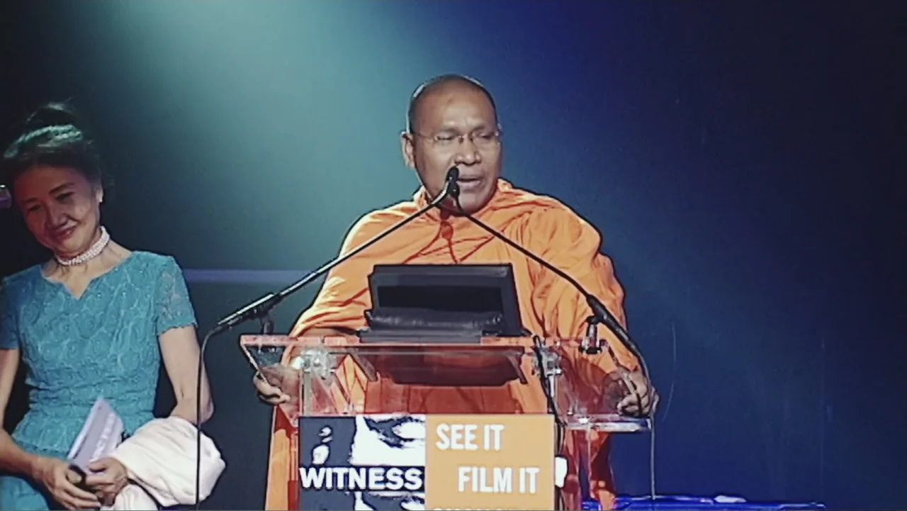 A Cambodian Spring (2018) movie still: medium — A man speaks at a podium with a 'Witness See It Film It' sign. A woman stands beside him holding…